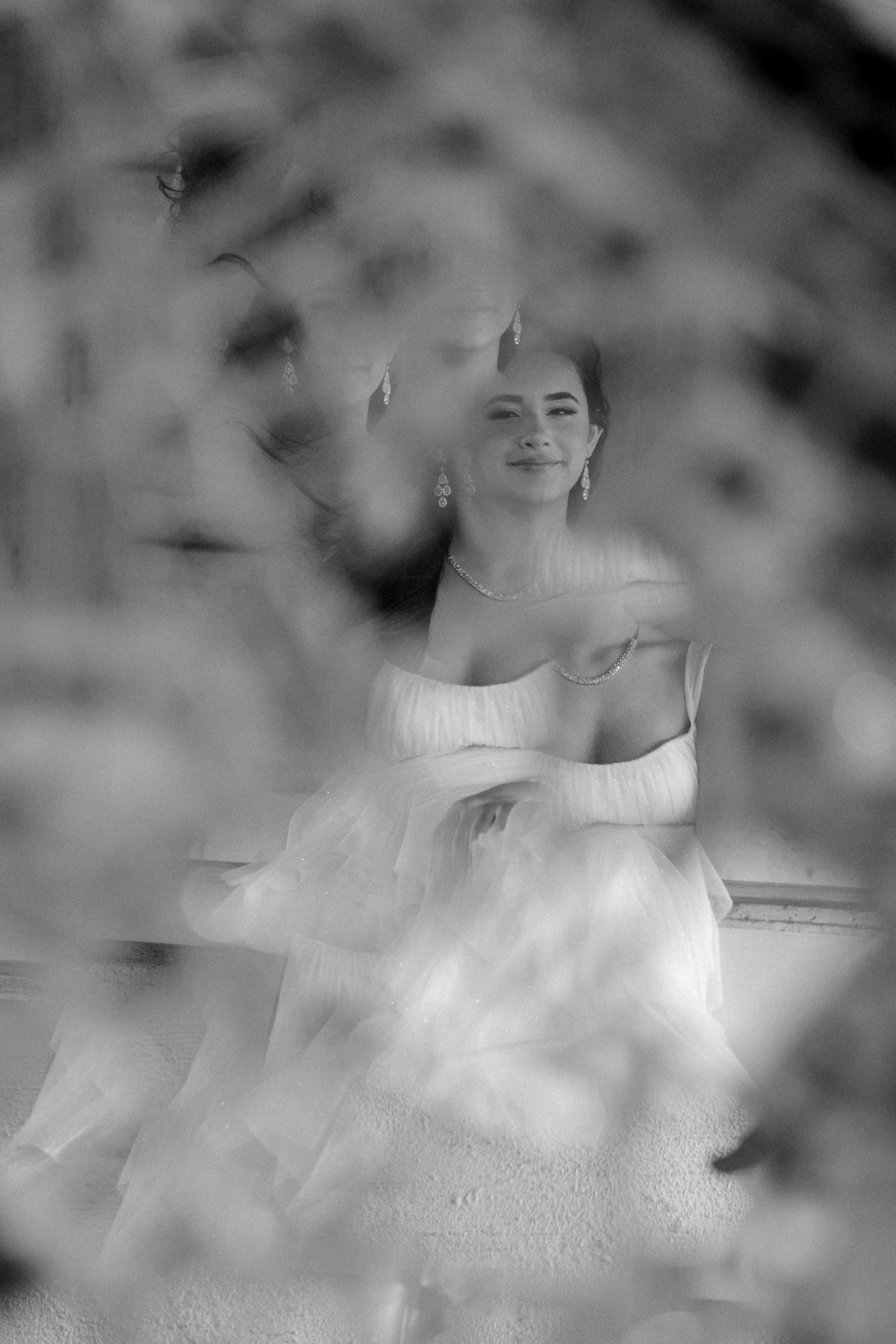 A dreamy black and white portrait of a Quinceañera in a white dress captured through blurred foreground elements.