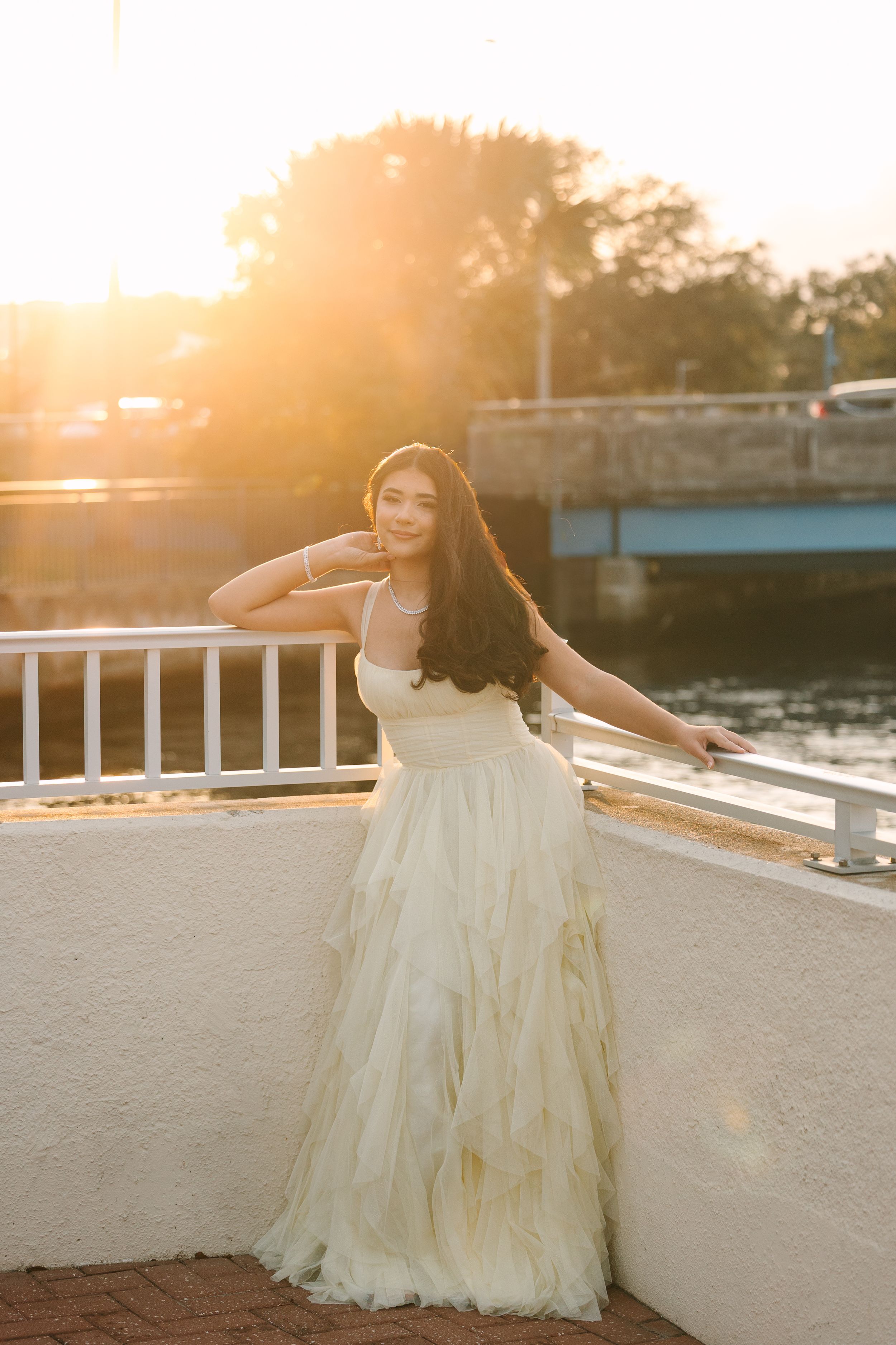 Quinceañera  in a flowing cream-colored gown poses gracefully against a sunset backdrop on a balcony.