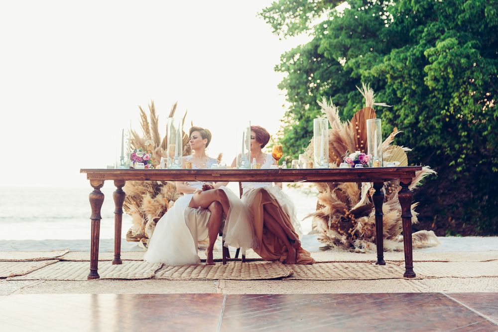 Elegant wooden table set for a wedding reception on a beach with dried palm fronds and natural decor.