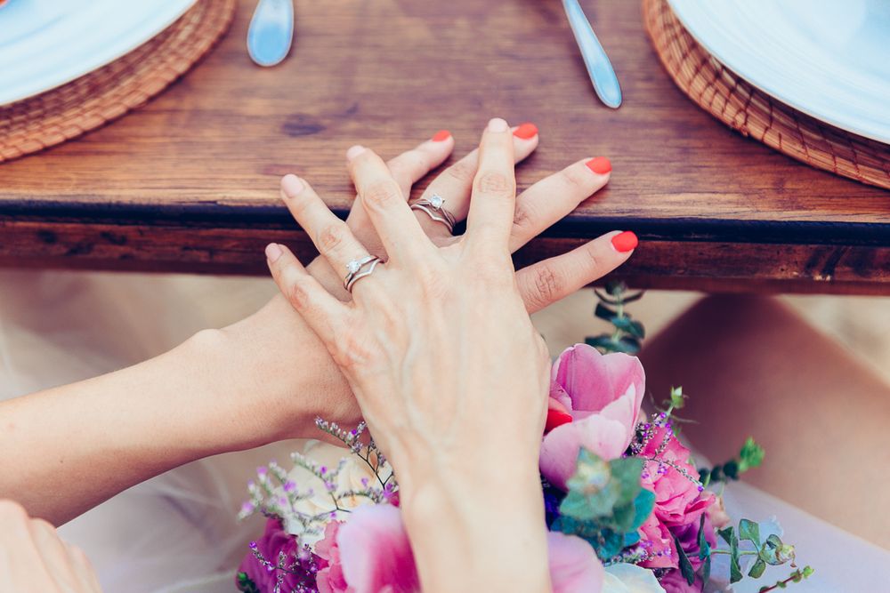 Hands displaying wedding rings among pink floral arrangements on wooden table.