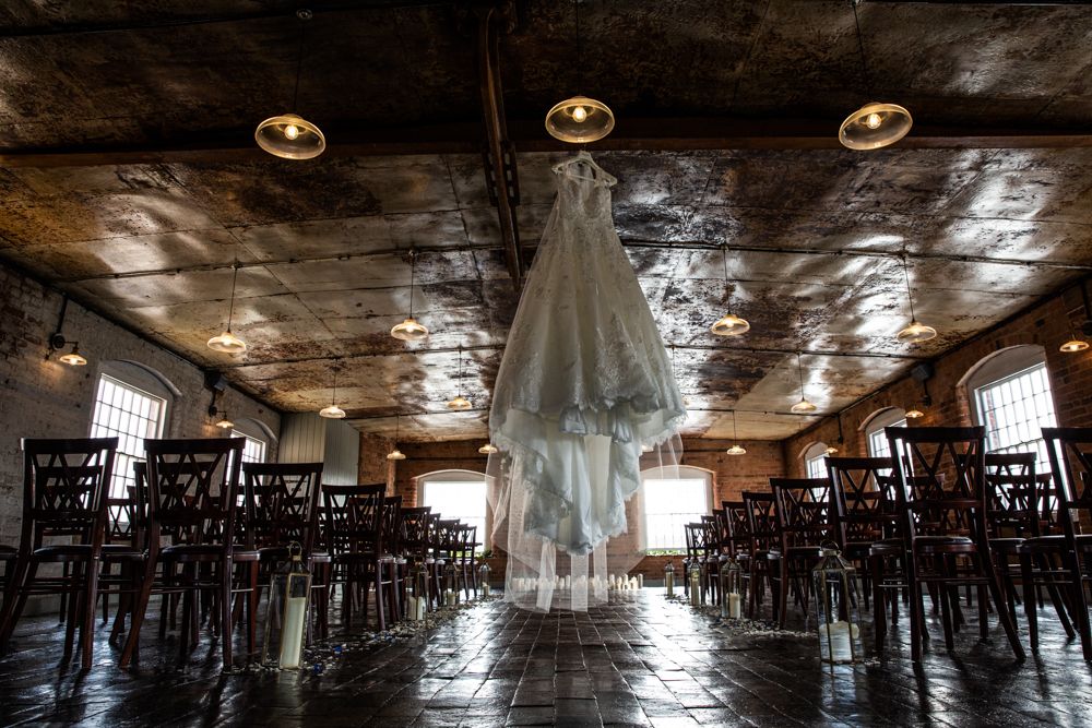 A wedding dress hangs in an empty rustic venue with wooden chairs and pendant lights.