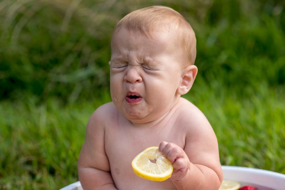 Funny baby making sour face while holding lemon slice sitting outdoors in white tub on grass.