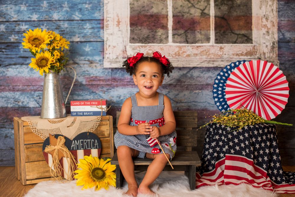 Toddler girl in patriotic American-themed photo session with sunflowers, flags, and red floral headband.