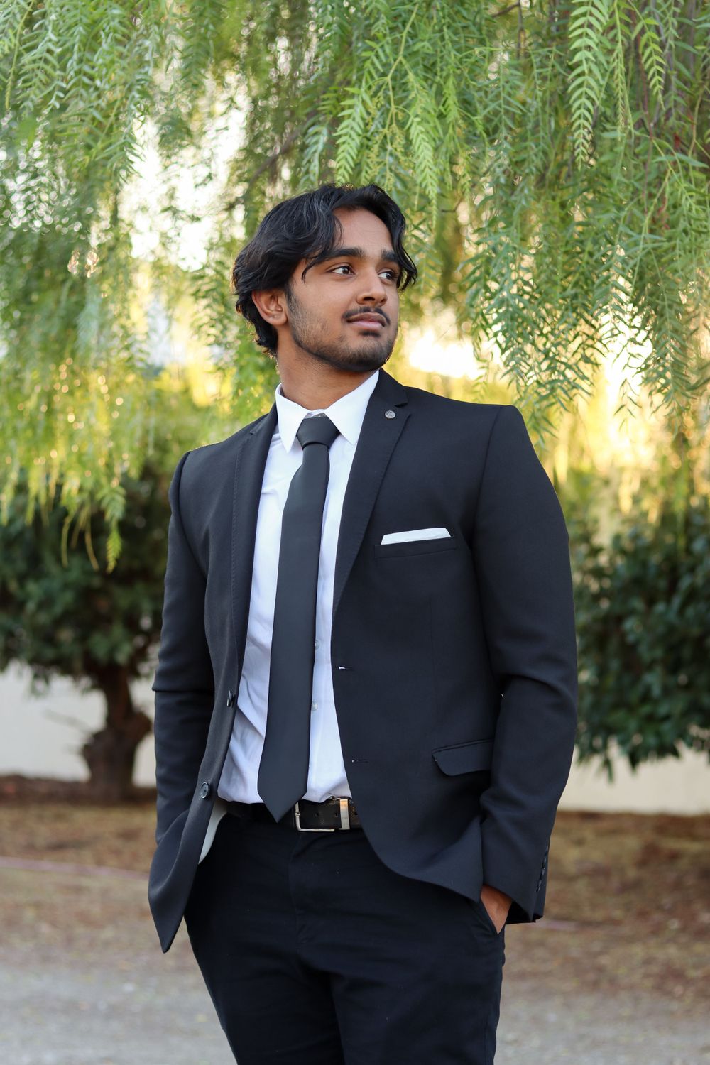 Man in black suit and tie poses confidently against outdoor garden setting.