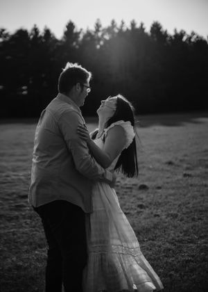 A romantic black and white portrait of a couple embracing in a field at sunset.