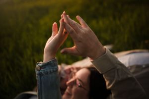 Hands reaching up towards sunlight in a grassy field during sunset.