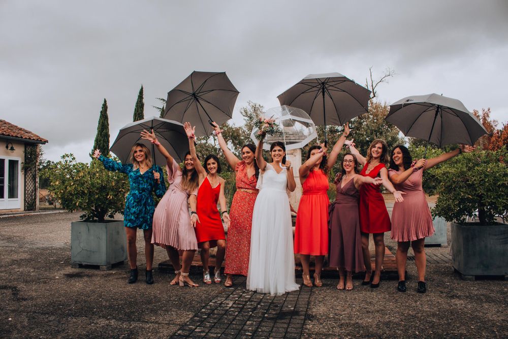 Wedding party with umbrellas posing joyfully in garden setting wearing vibrant dresses.