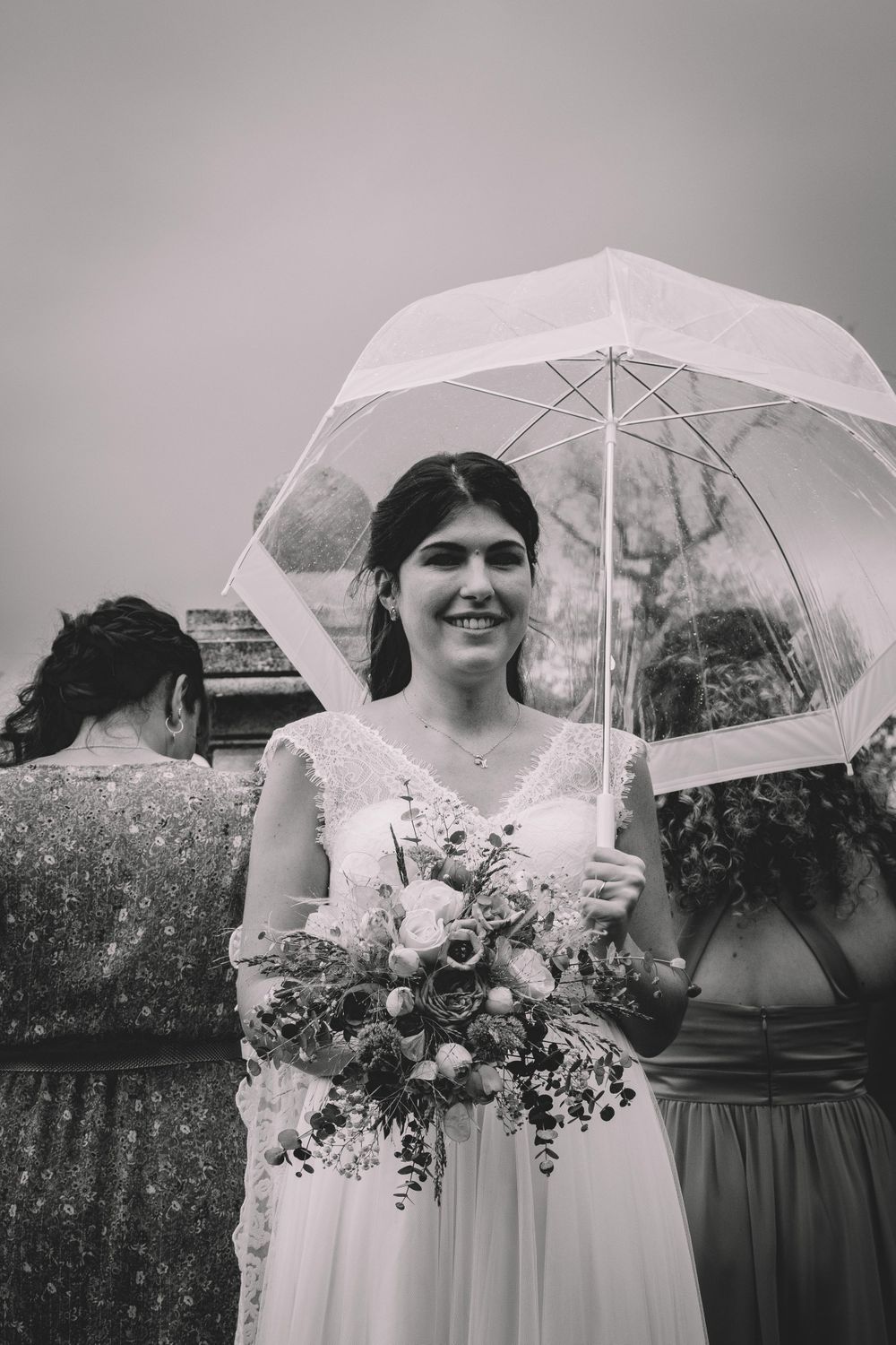 A bride in white dress holds a clear umbrella while carrying a floral bouquet.