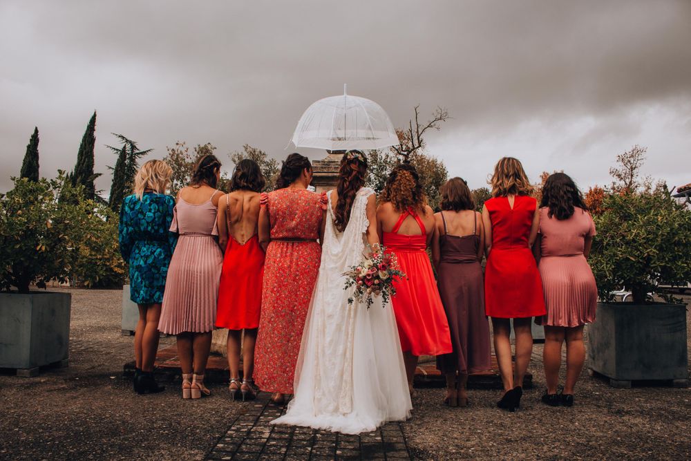 Wedding party facing away showing back details of colorful dresses against cloudy sky.