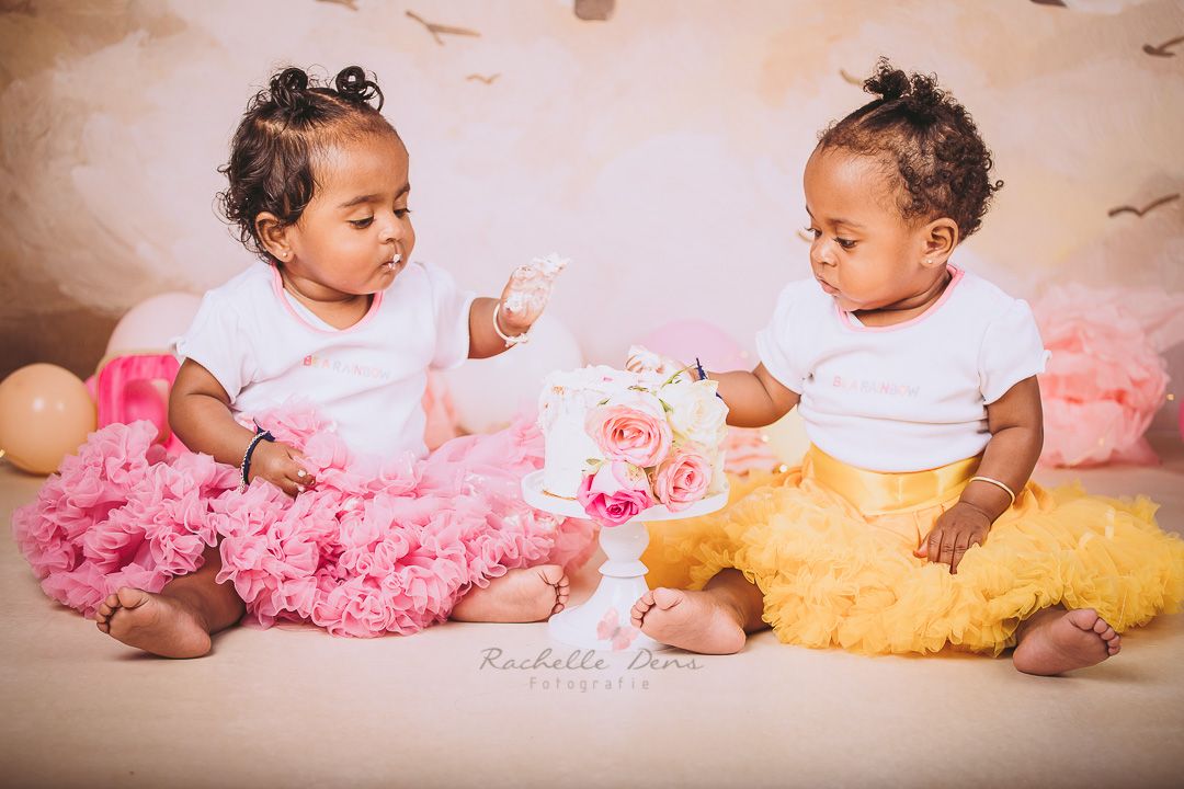 Twin babies in matching white tops with pink and yellow tutus lying on white backdrop.