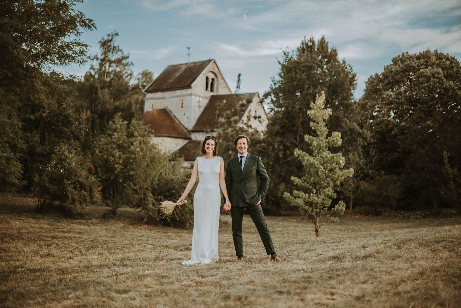 Couple in wedding attire poses together on grassy field with farmhouse in background.