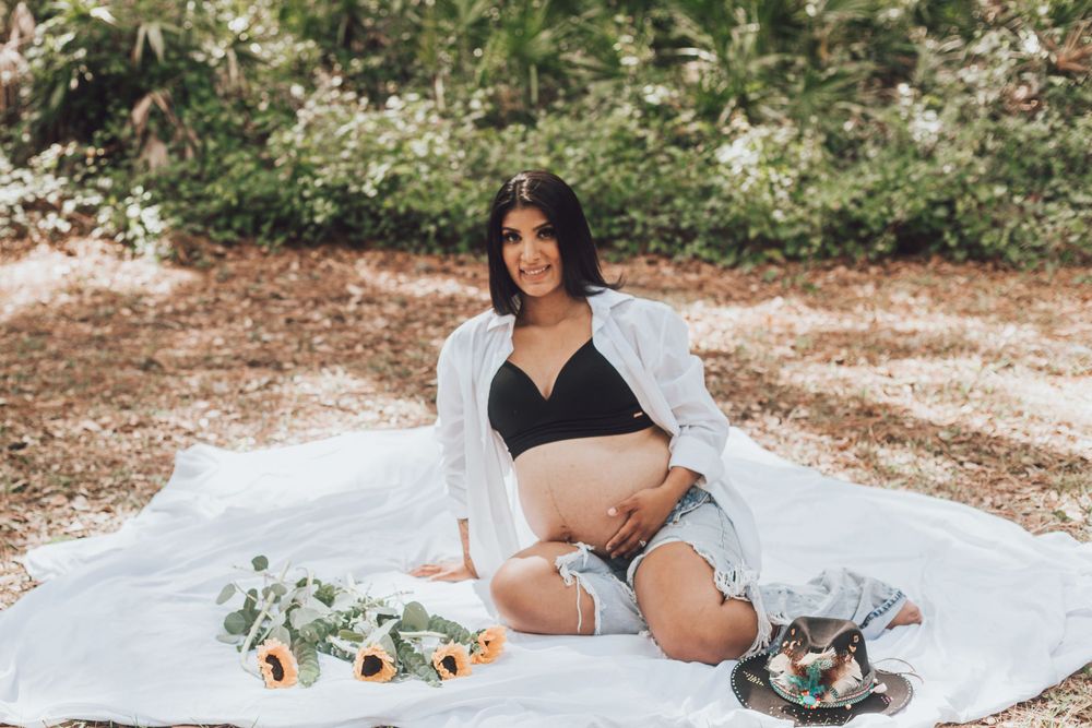 Maternity photo of an expectant mother in black lingerie sitting on white blanket outdoors.