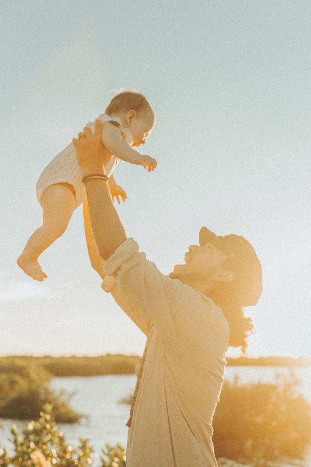 A parent lifting a baby against a golden sunset backdrop near a waterfront.