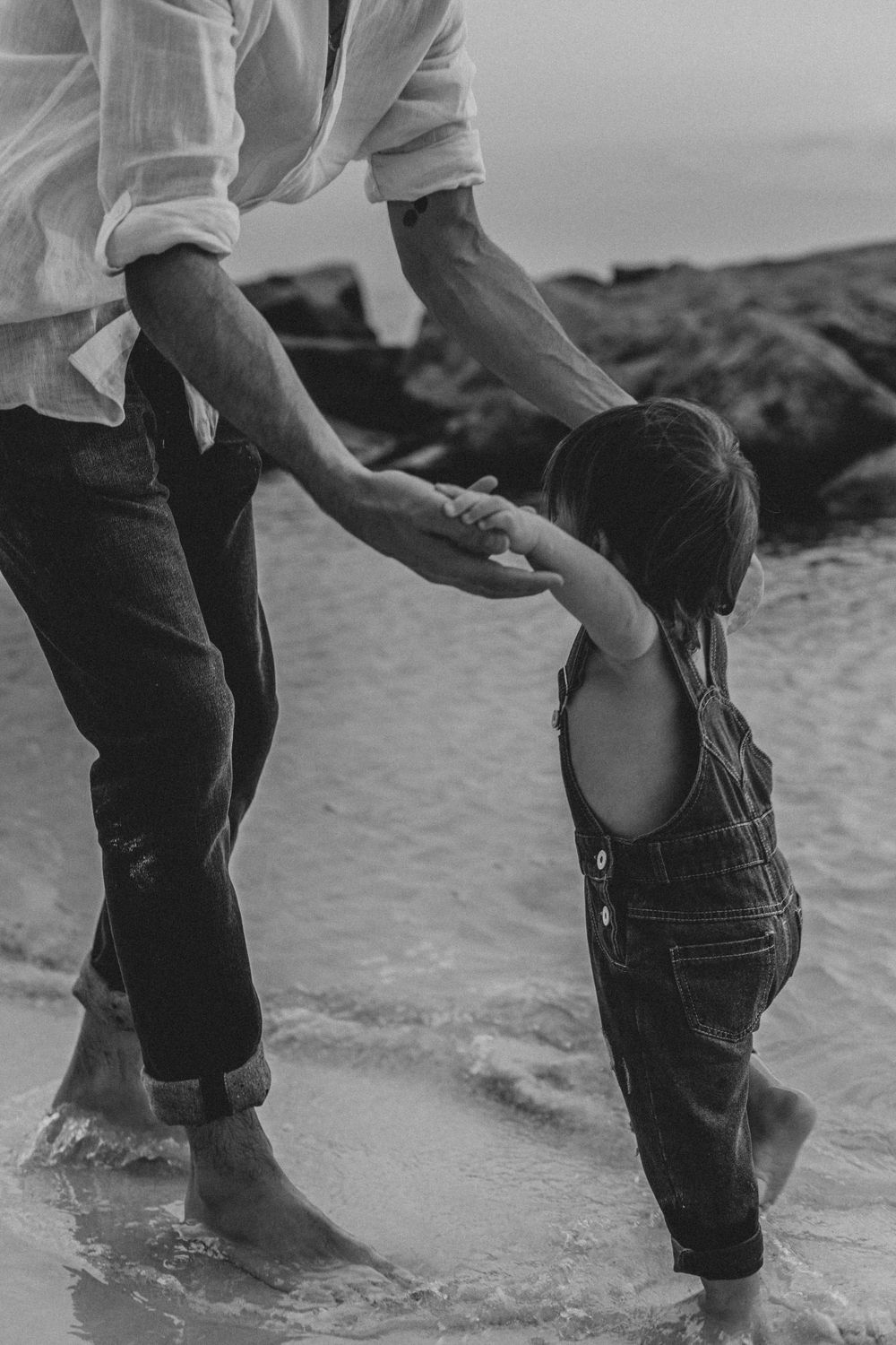 Parent and child hold hands while standing in shallow ocean water, captured in black and white.