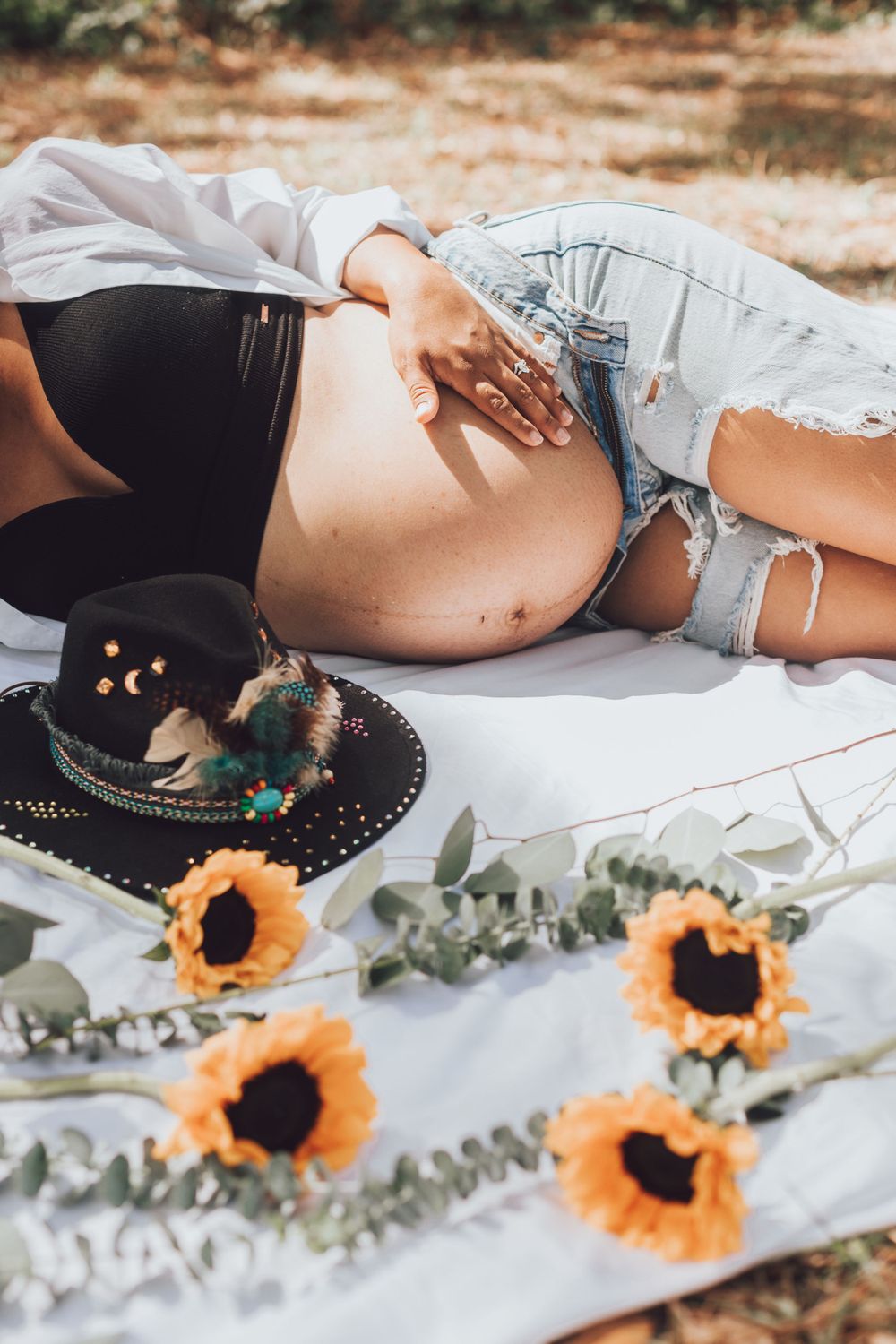 Artistic maternity photo with sunflowers and black hat on white blanket outdoors.