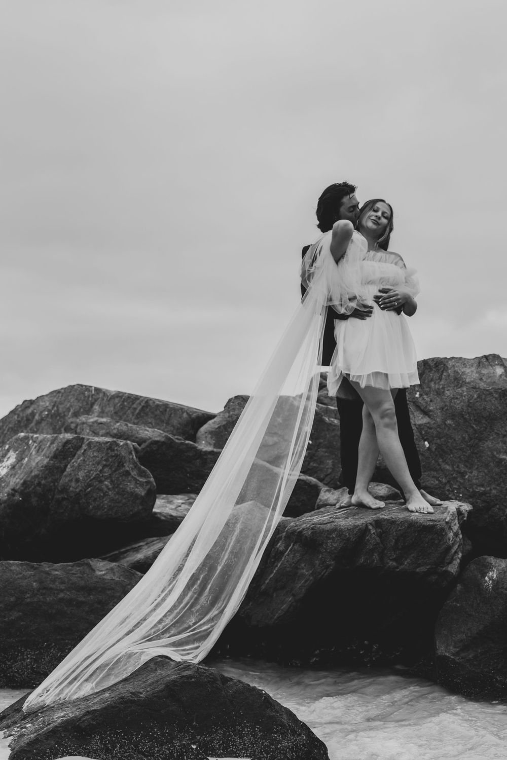 Black and white beach portrait with flowing veil against dramatic rocky backdrop.