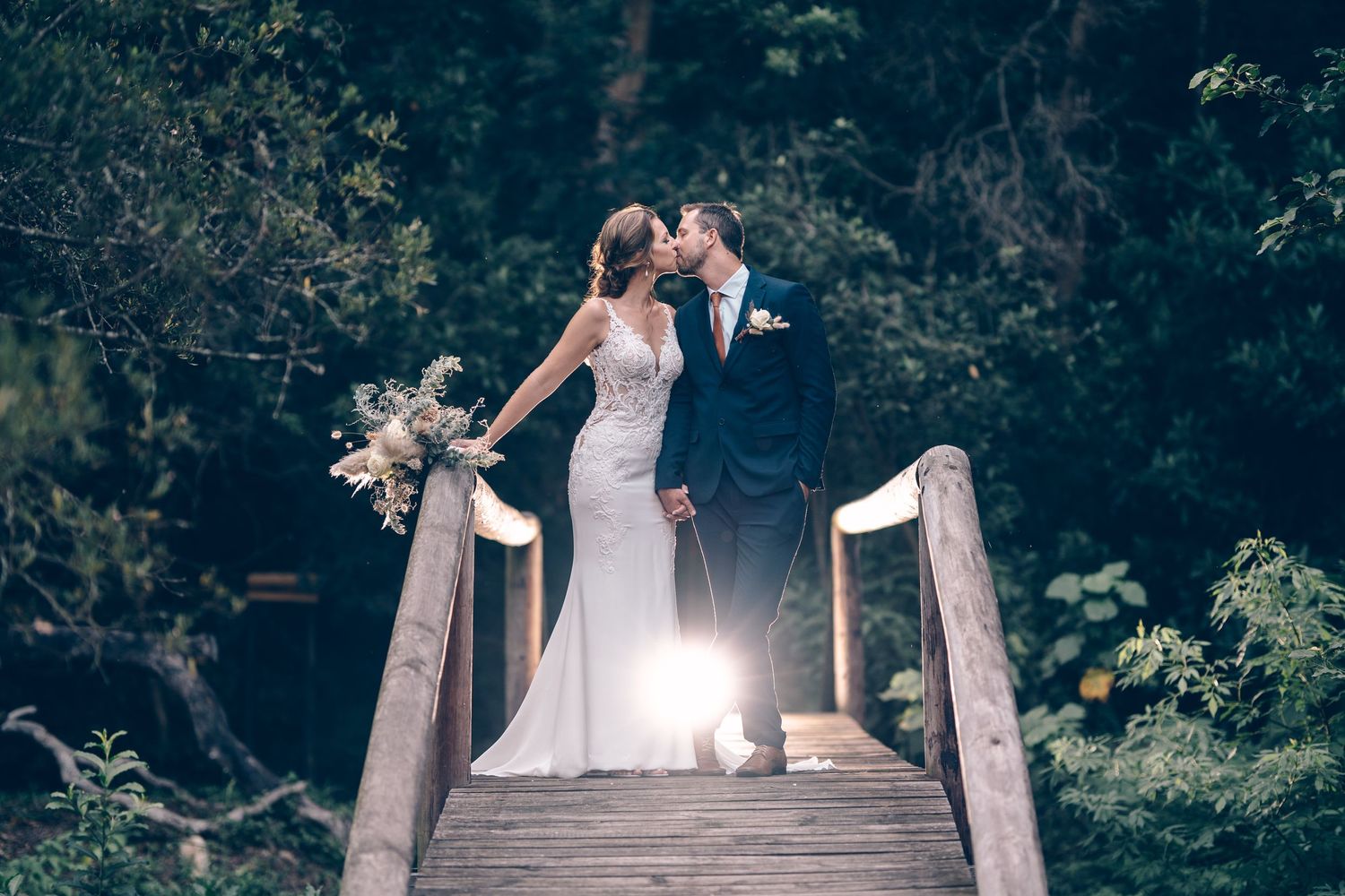 Bride and groom sharing a kiss on a wooden bridge surrounded by lush green forest at night.