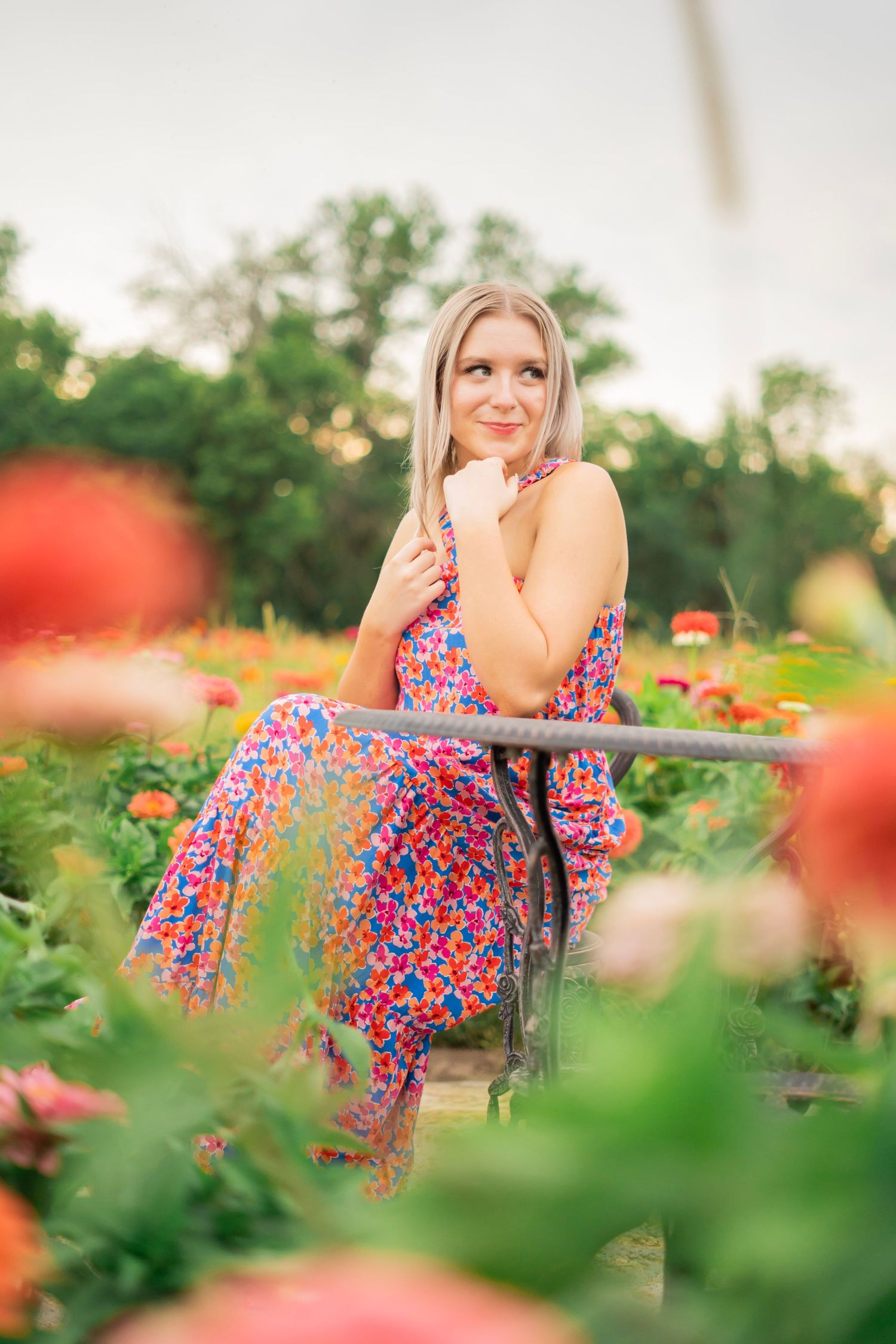 A blonde woman in a colorful floral dress sits on a bench surrounded by vibrant zinnia flowers at sunset.