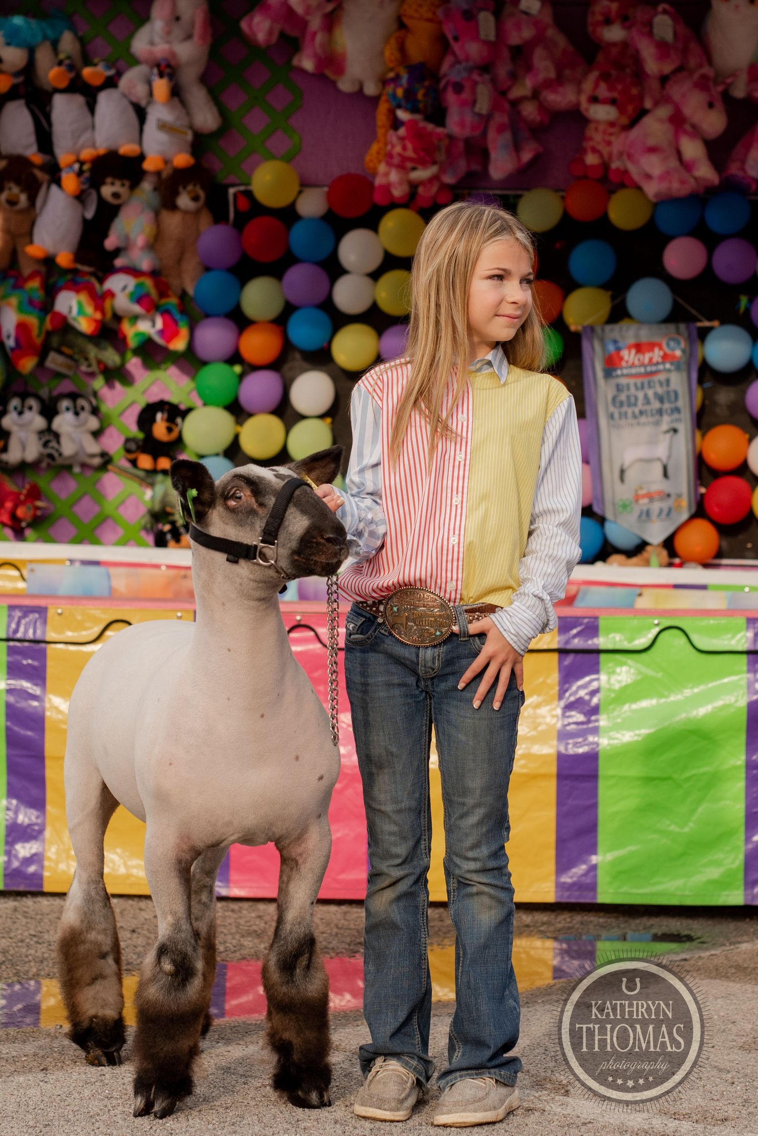 A contestant stands with a show sheep at a colorful county fair carnival booth with stuffed animals and balloons.