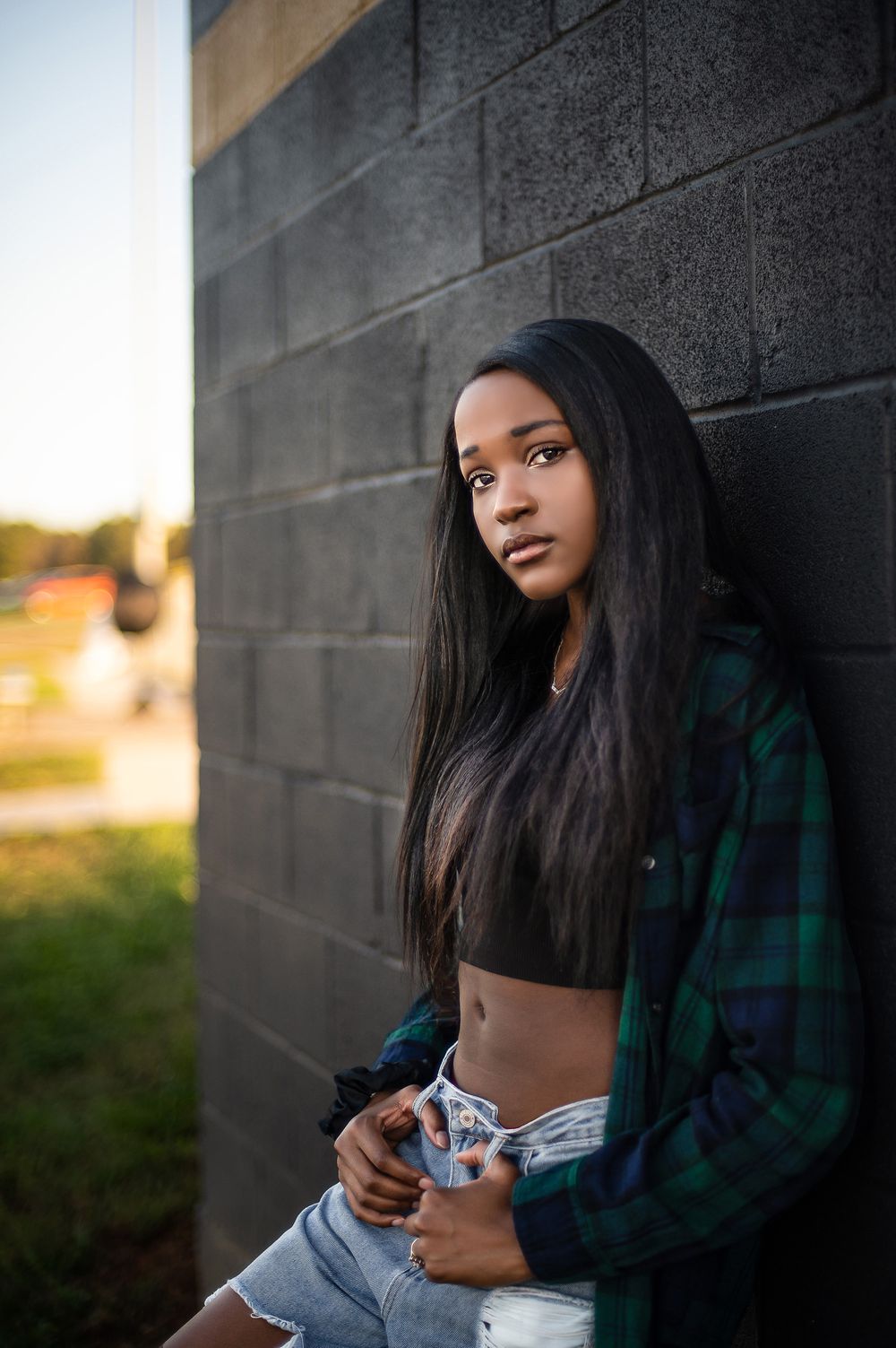 Woman in plaid jacket poses against dark wall at sunset with dramatic lighting and shadows.