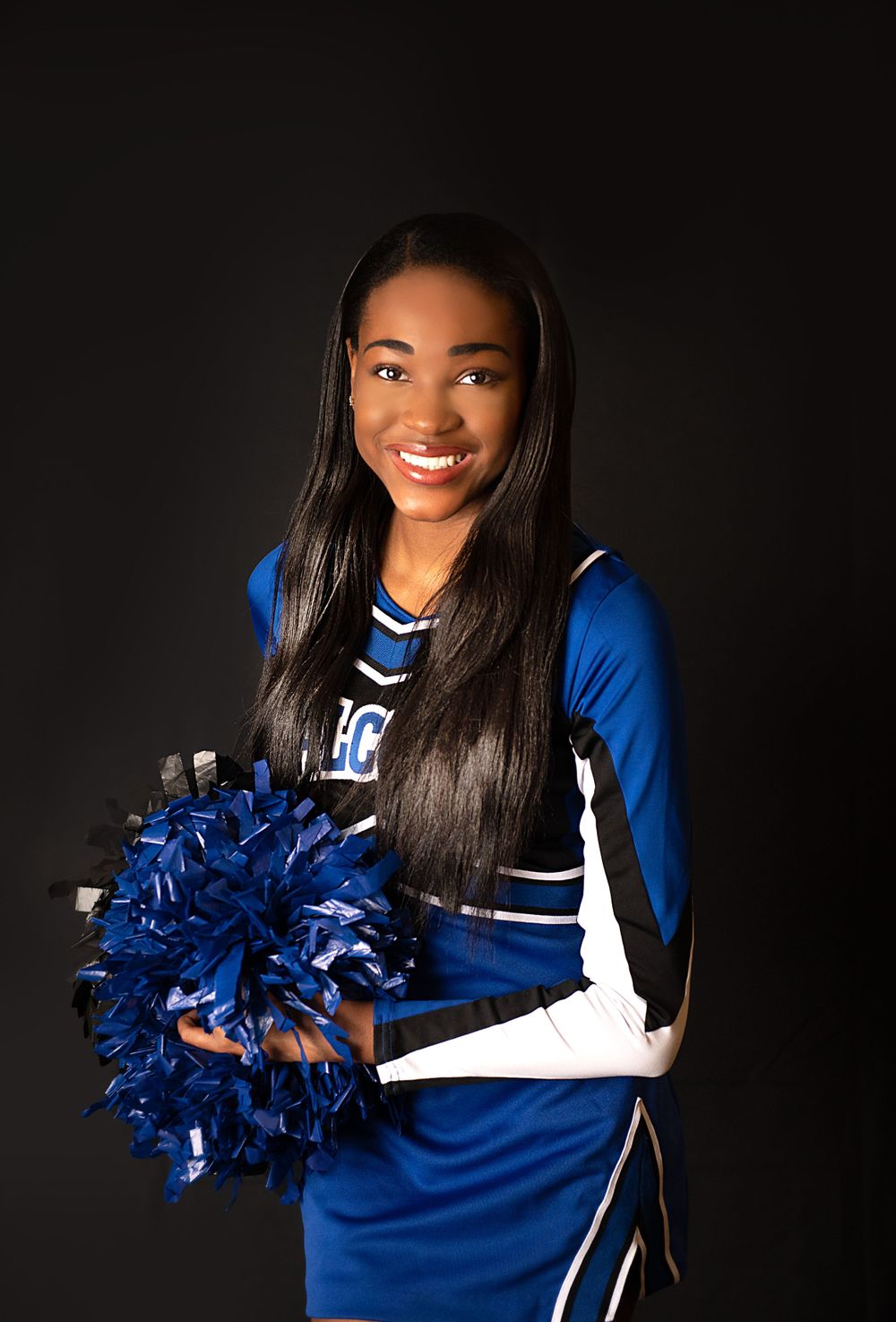 High school cheerleader in blue and white uniform holds blue pom poms against dark background.