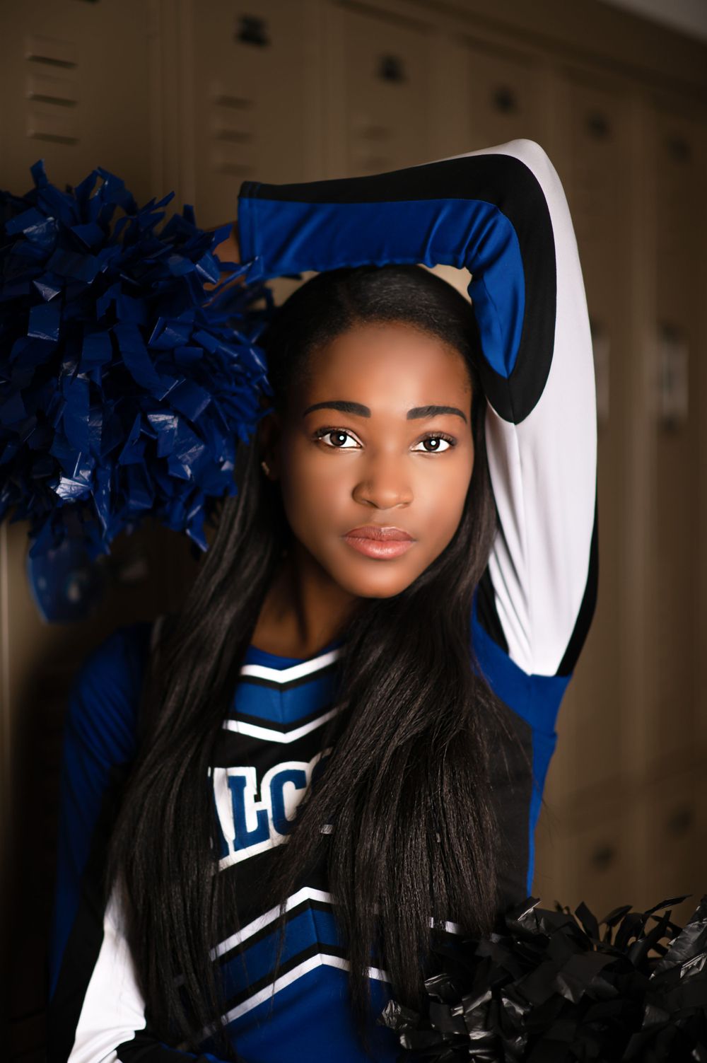 Cheerleader in blue uniform poses with pom poms against indoor background.