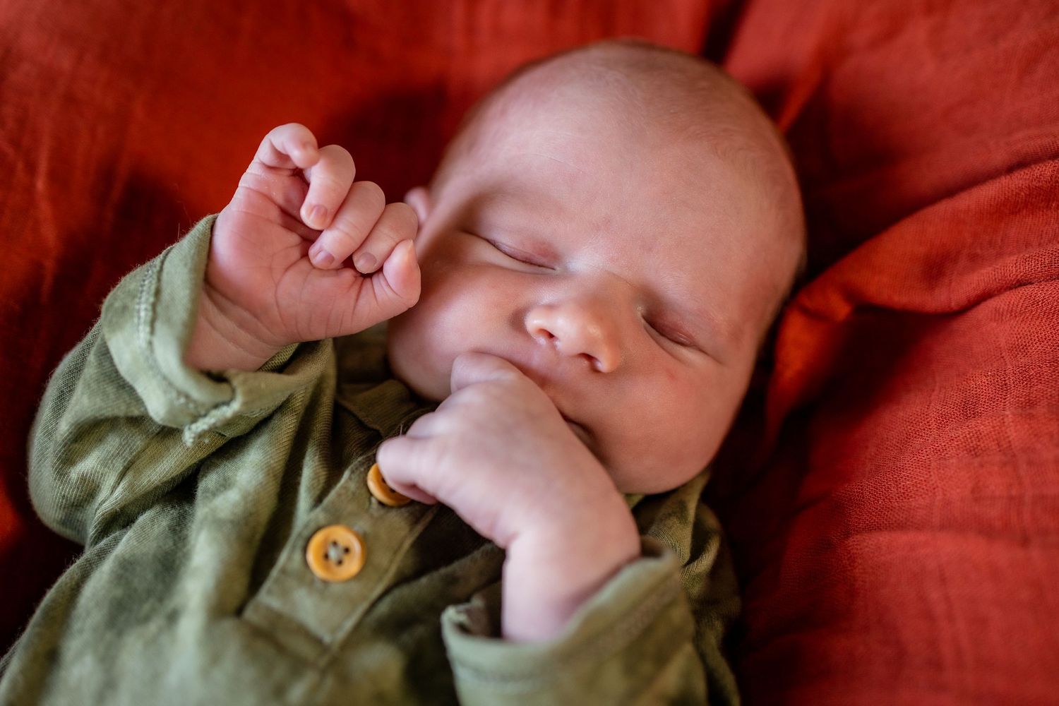 Peaceful newborn baby sleeping in green outfit on red blanket with tiny fist raised.