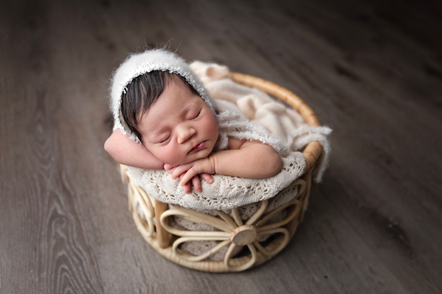 Sleeping newborn with white headband posed in woven basket on wooden floor.