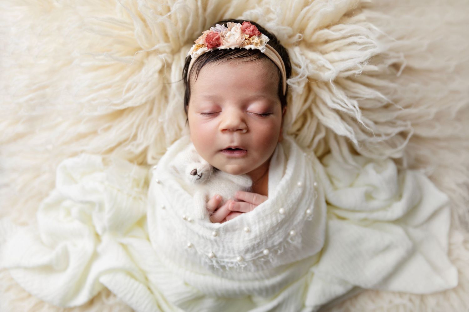 Newborn wrapped in white lace blanket on cream textured backdrop with floral headband.