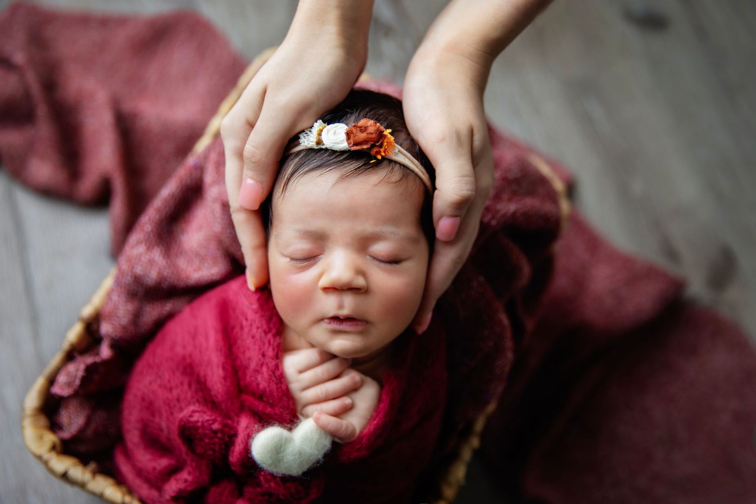 Sleeping newborn in red wrap and headband on burgundy blanket.