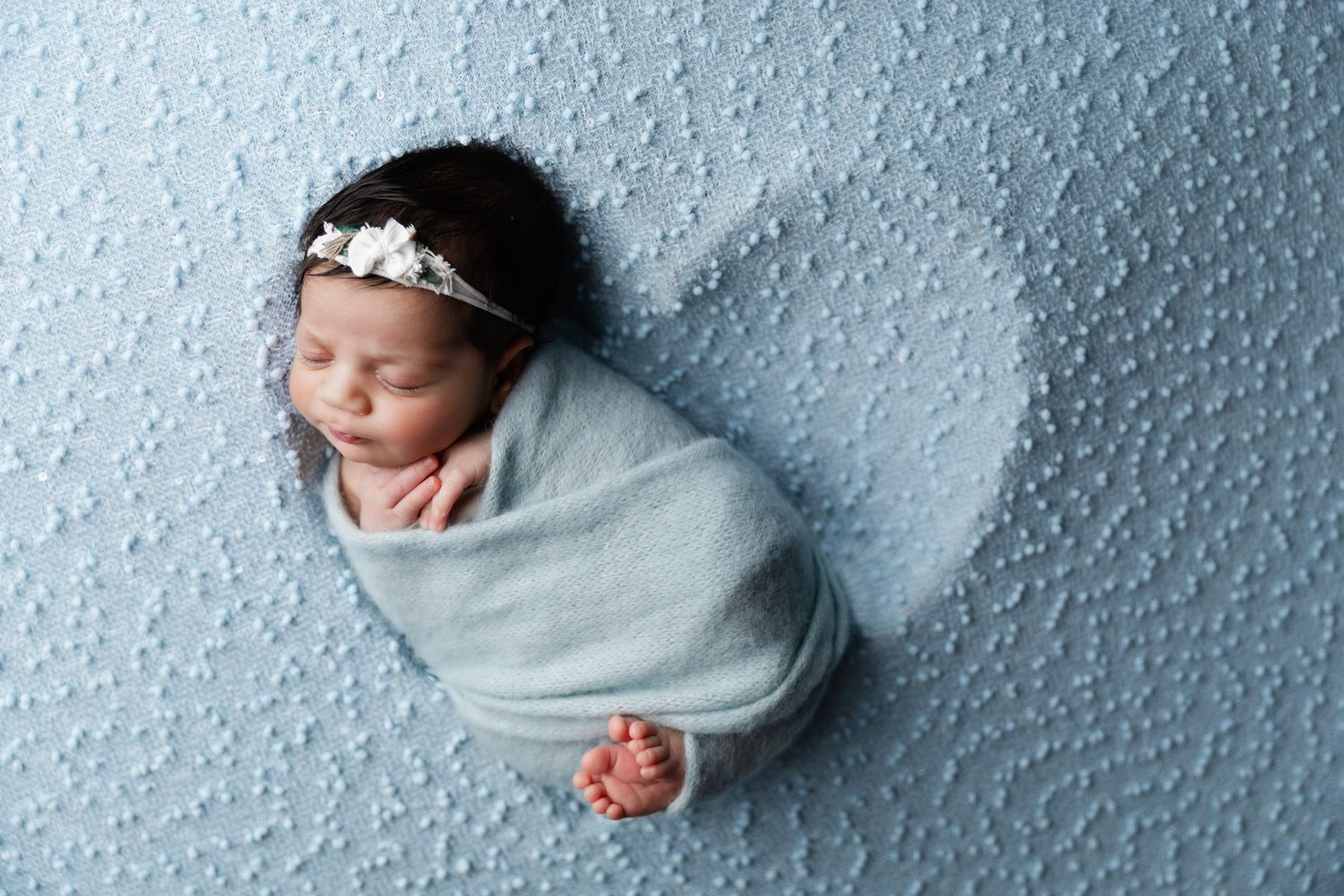 Peaceful newborn wrapped in gray on textured blue background with white headband.