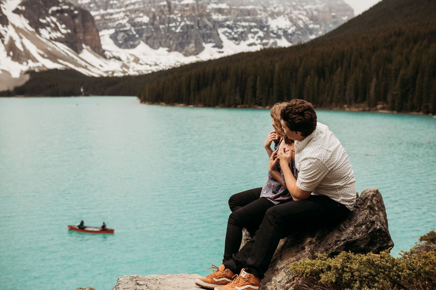 Person sitting on rock overlooking turquoise alpine lake with snowy mountains in background.