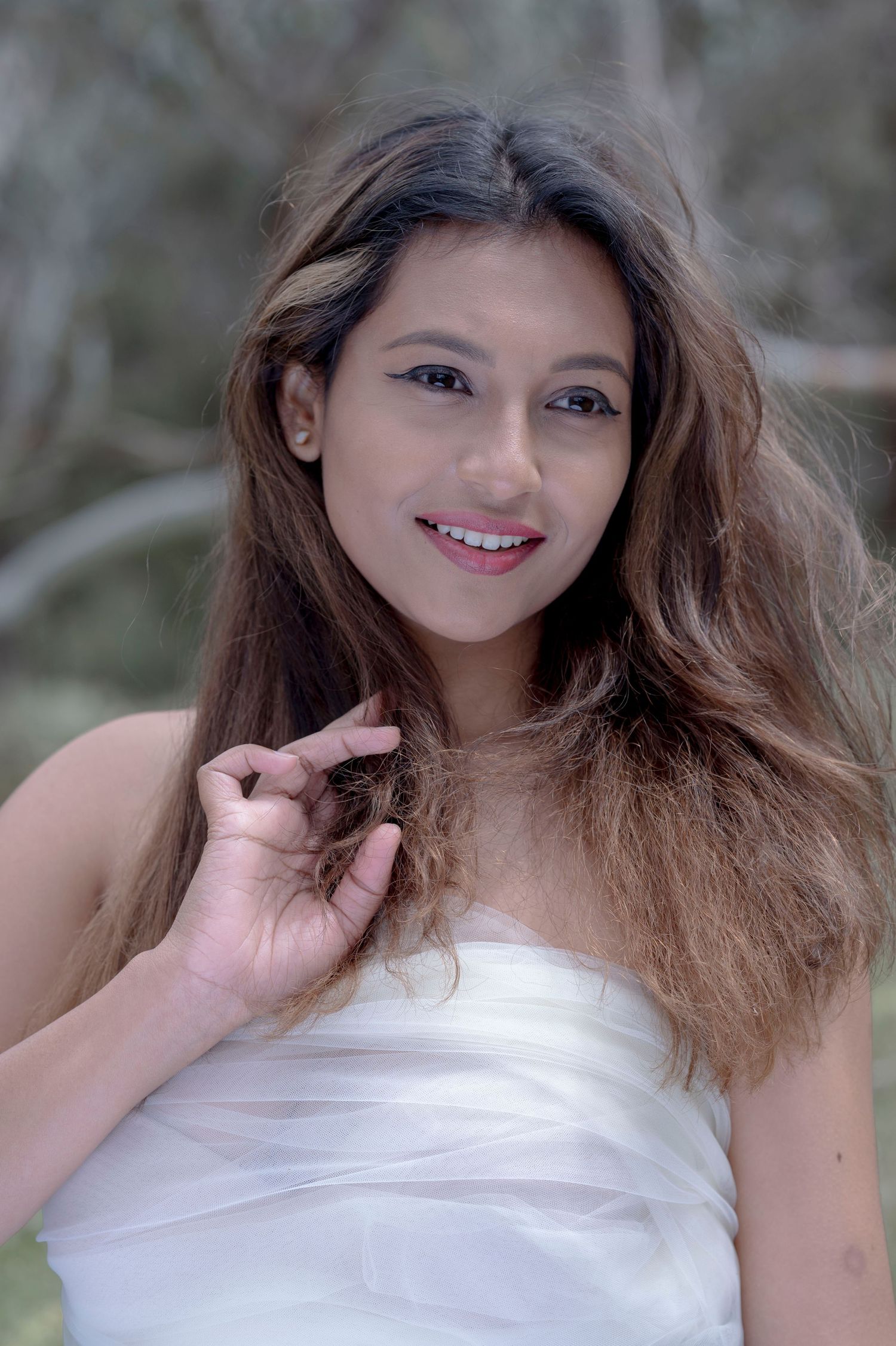 Smiling young woman with long brown hair wearing a white strapless top poses outdoors.
