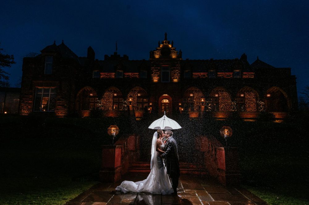 A couple shares an umbrella in front of an illuminated historic building at night during their wedding.