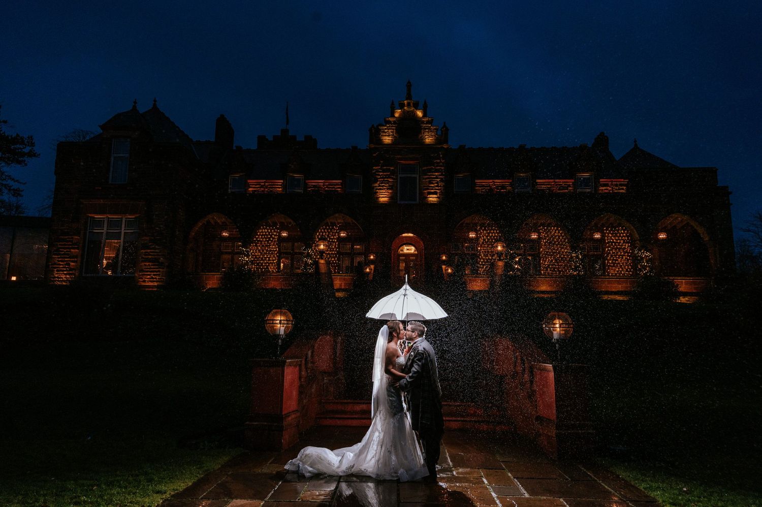 A couple shares an umbrella in front of an illuminated historic building at night during their wedding.