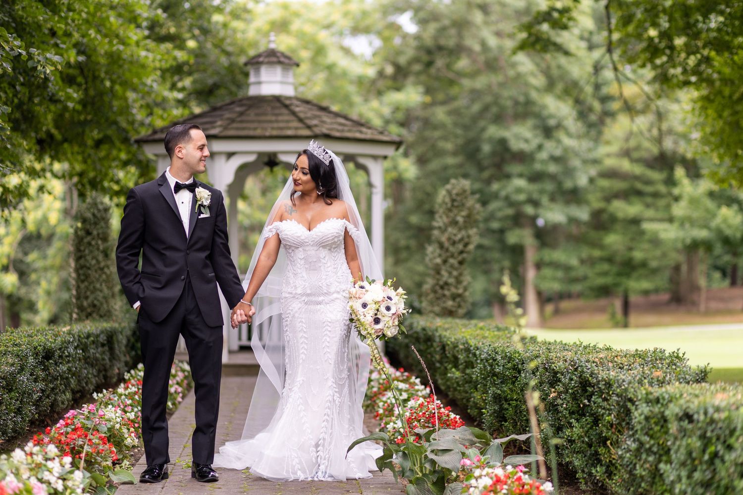 A bride and groom pose in a garden setting with a white gazebo and colorful flowers.