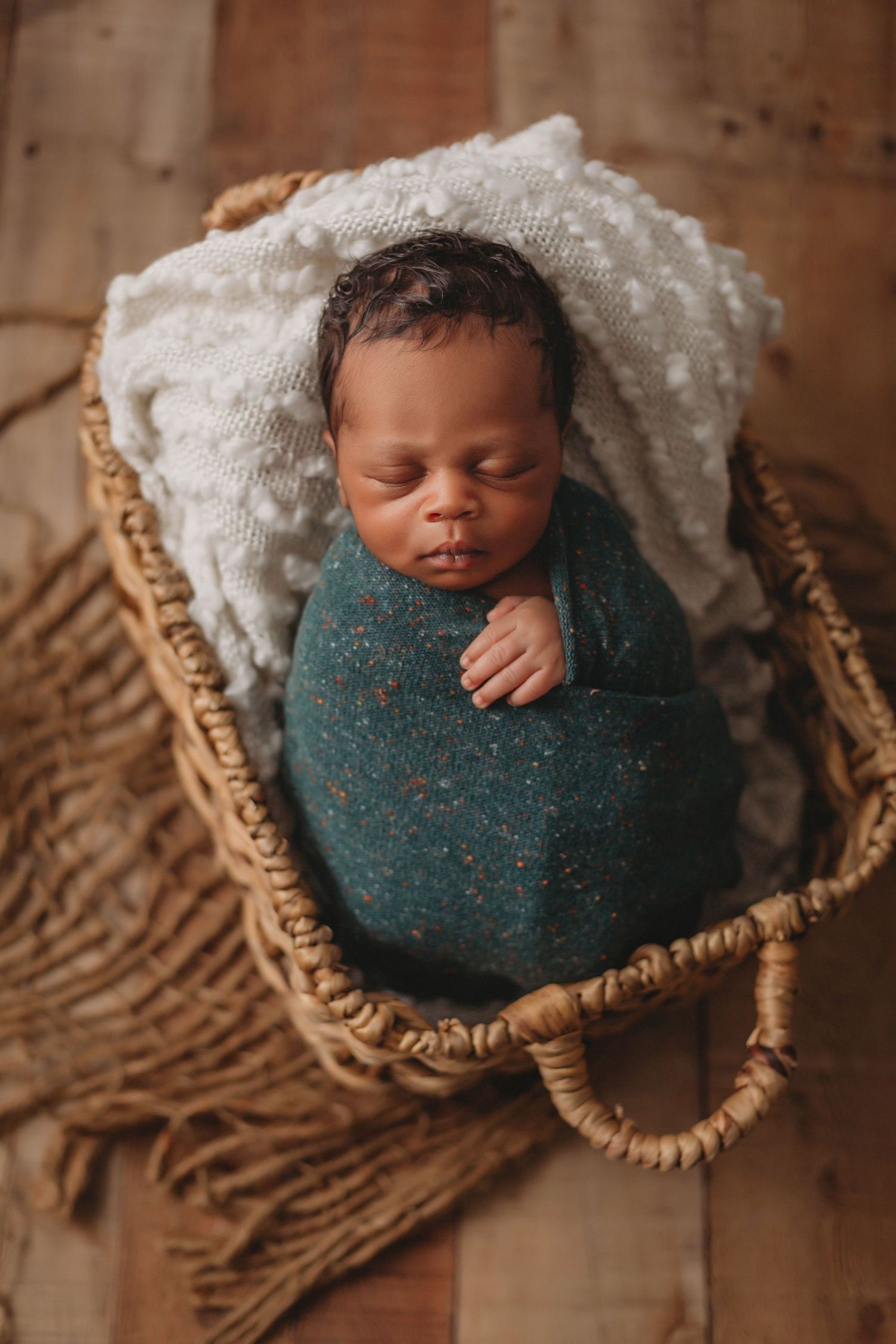 Sleeping newborn wrapped in teal blanket resting in woven basket on wooden floor.