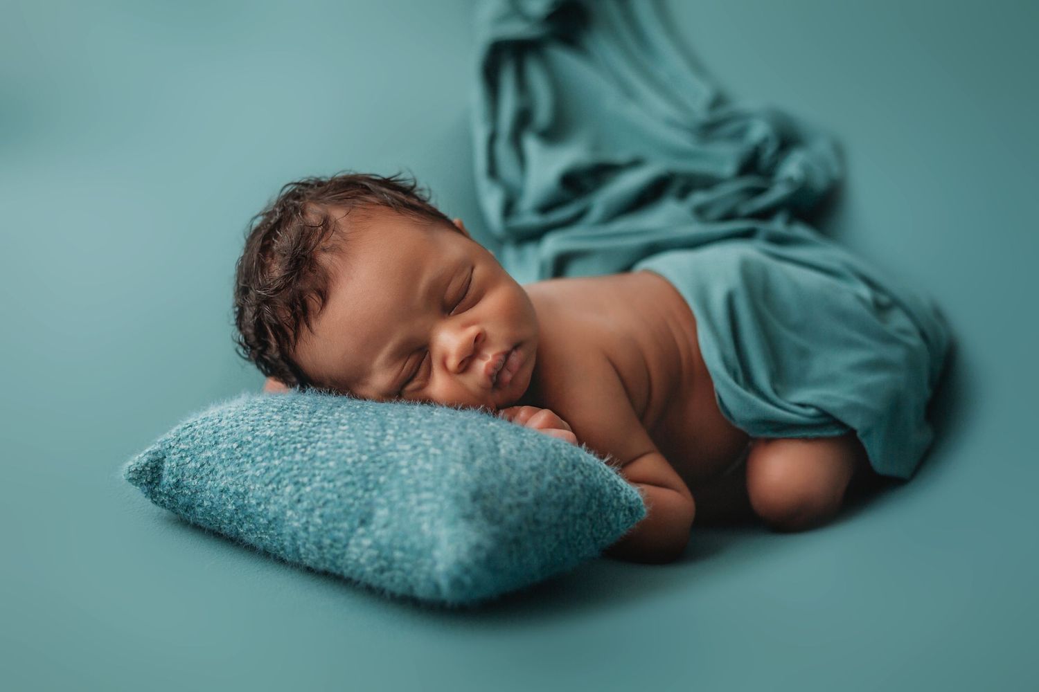 Peaceful sleeping newborn wrapped in teal fabric on matching blue pillow.
