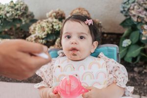 Floral-dressed baby sits among decorative flowers and pink balloons at a celebration.