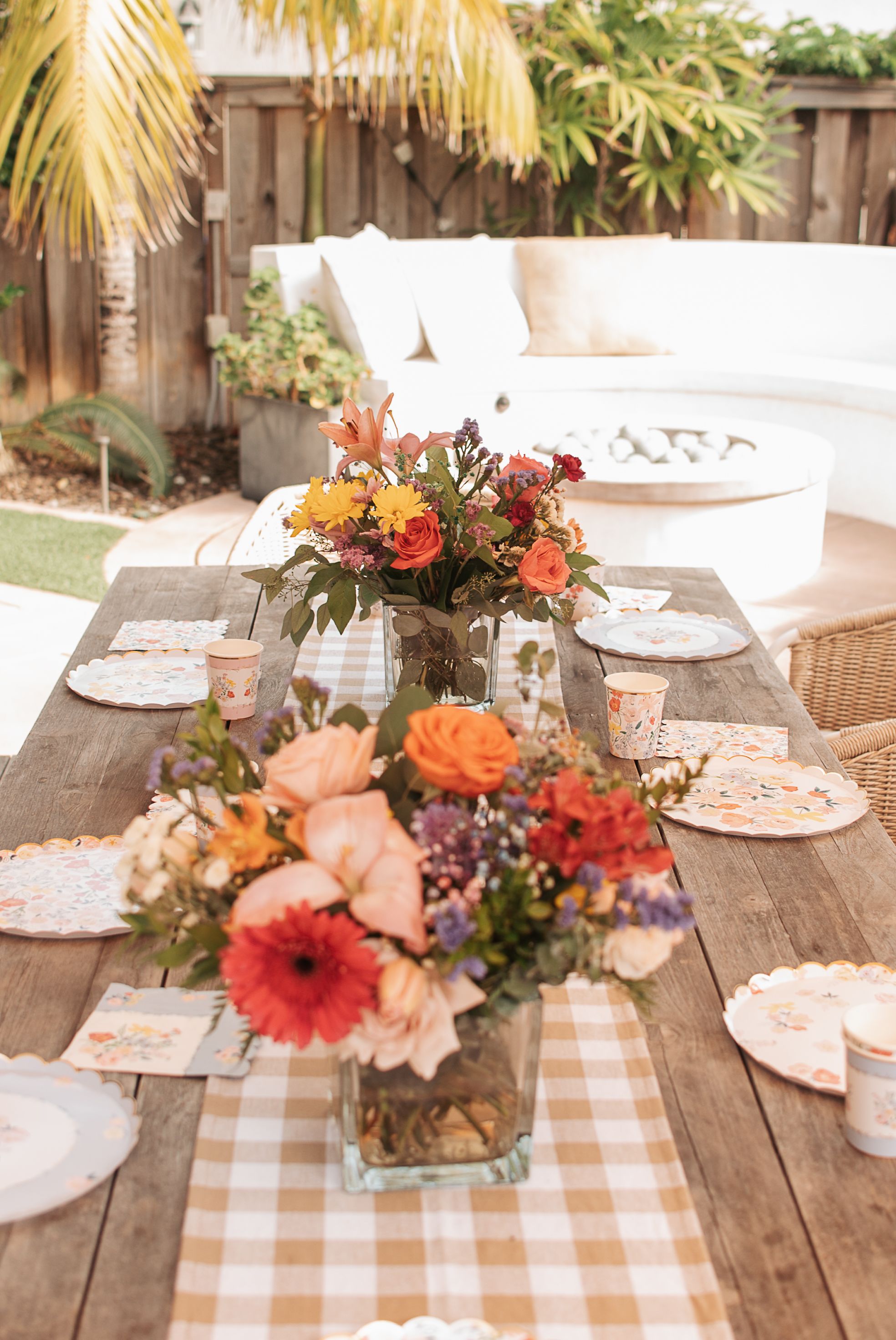 Outdoor table setting with gingham runner and colorful floral centerpieces on rustic wooden surface.