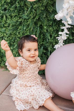 Smiling baby in lace dress plays near pastel balloons and garden foliage.
