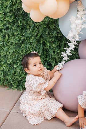 A baby in white dress plays near peach balloons and greenery at an outdoor party setting.