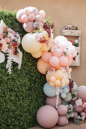 Pastel balloon arch with pink and peach balloons decorated with fresh flowers against a garden wall.