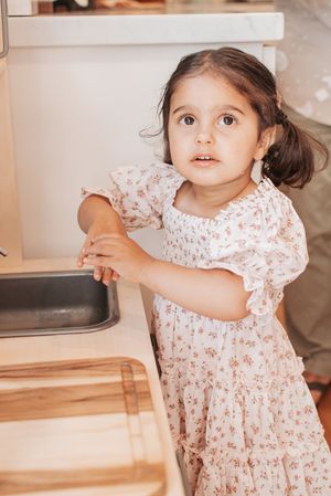 A toddler in a floral dress plays around a wooden kitchen setup with a curious expression.