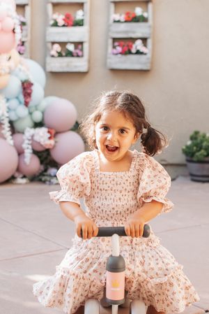Young child in a ruffled white dress twirls joyfully in front of a decorated wall with balloons.