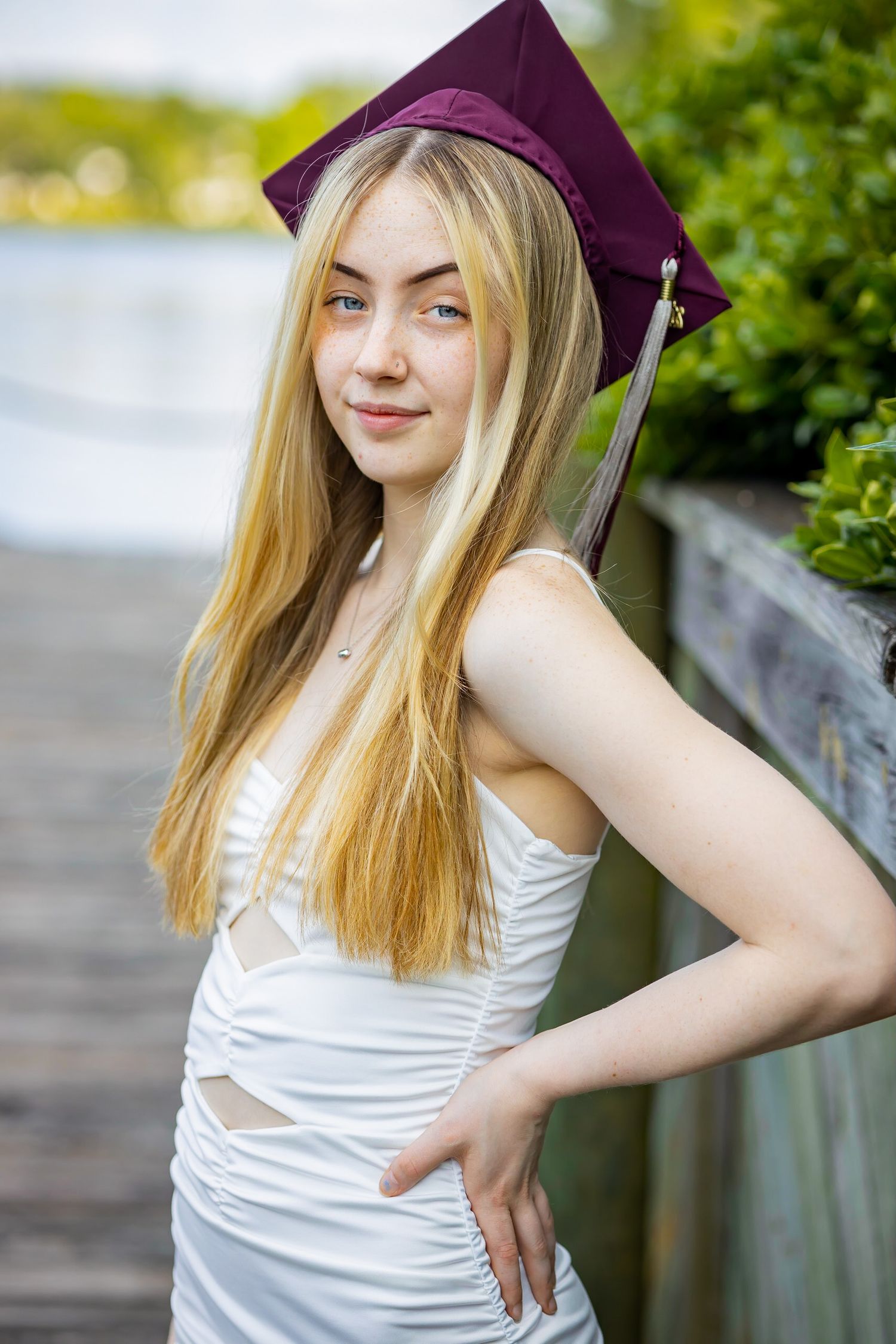 A graduate in maroon cap and white dress poses casually against railing during photo session.
