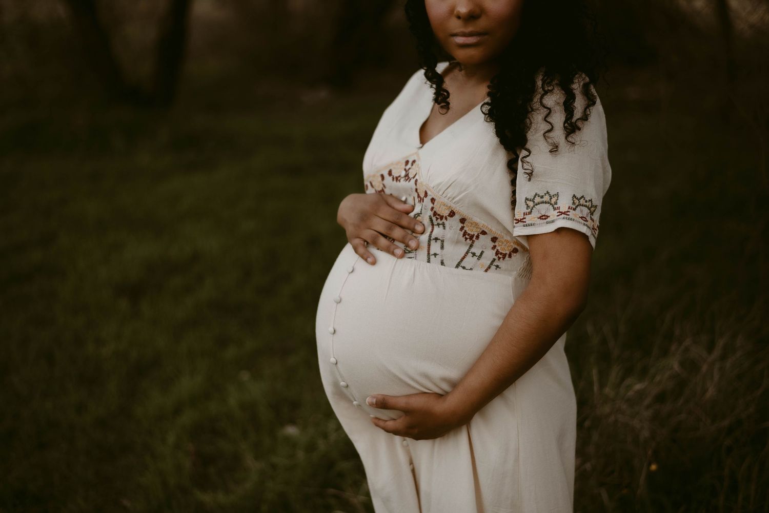 Side profile maternity photo showing a white dress with delicate embroidery details against darkened background.