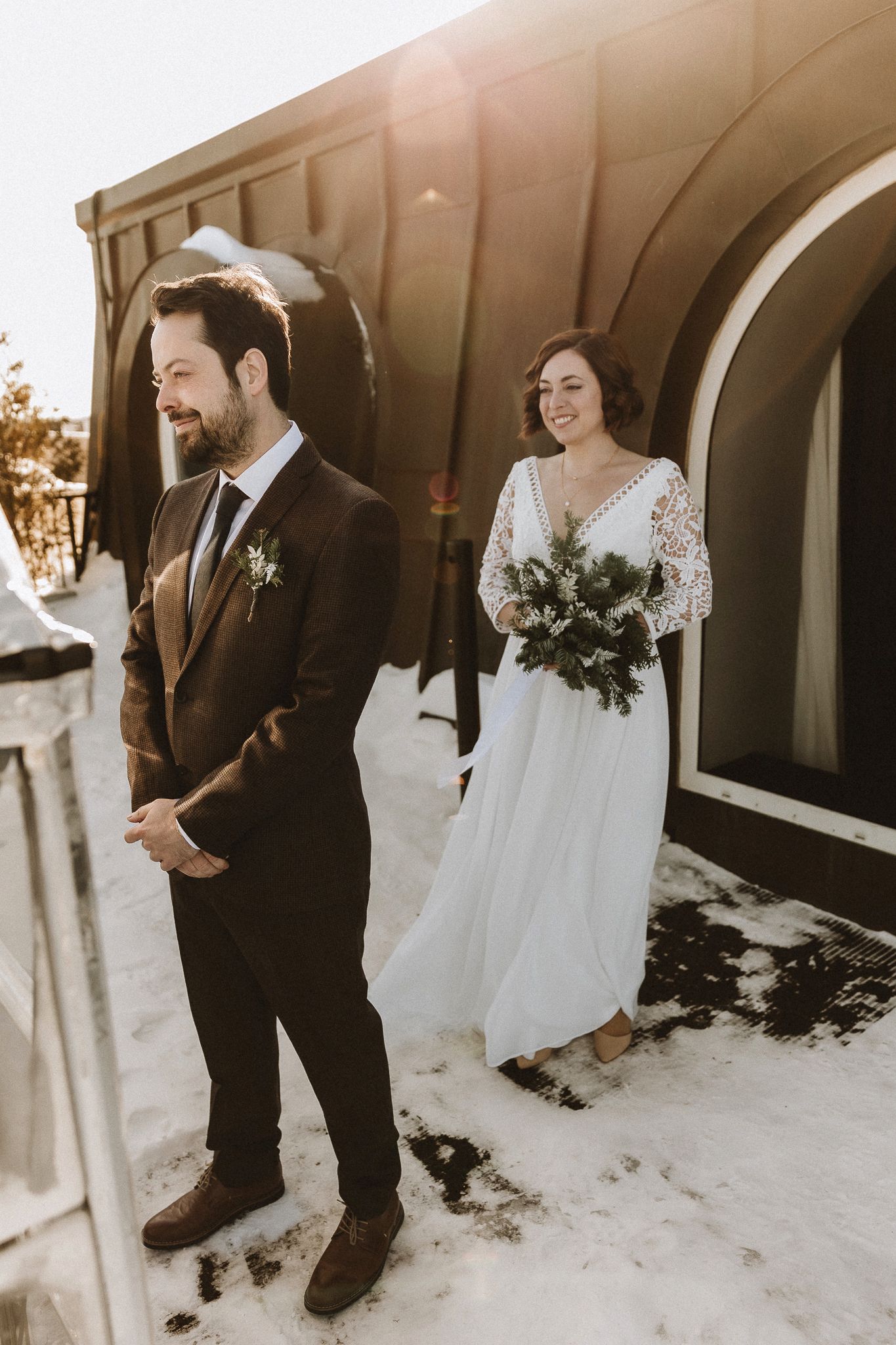 A winter wedding first look moment against snowy ground and an arched building exterior.