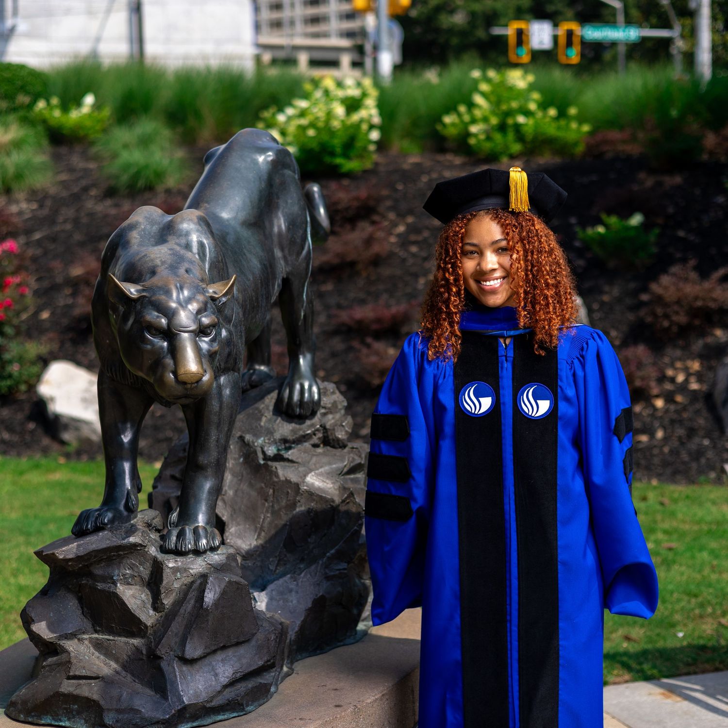 A majestic black panther statue stands next to a graduate in blue academic regalia on a university campus.