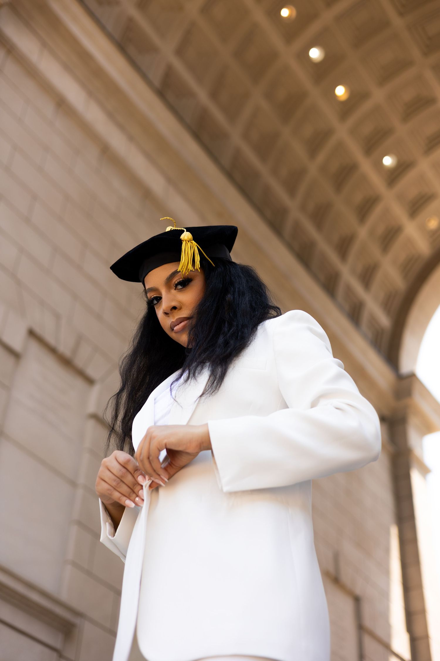 Graduate in white blazer and cap photographed from below against architectural ceiling.